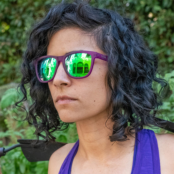 Person wearing Purple Goodr OG Gardening With a Kraken sunglasses with blue lenses standing outdoors with greenery in the background