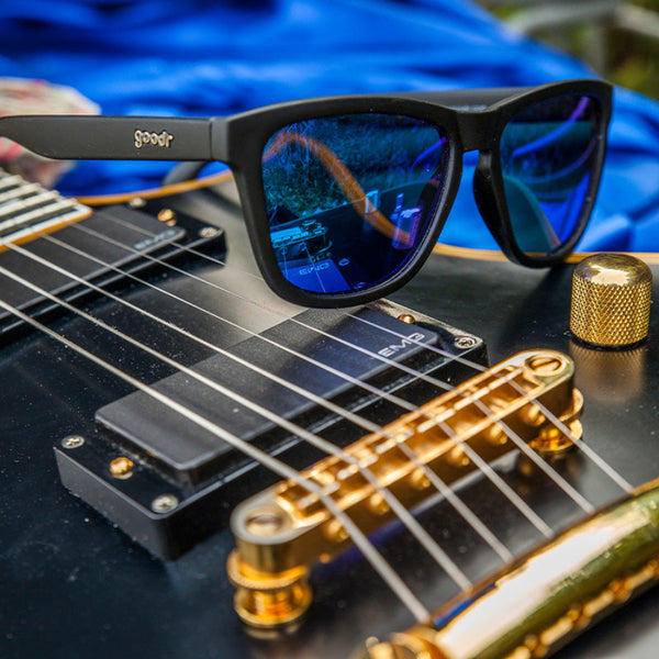 Goodr OG Sunglasses with blue lenses on a black electric guitar with gold hardware.