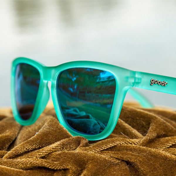 Turquoise Goodr OG sunglasses with green lenses on a textured surface and neutral background.