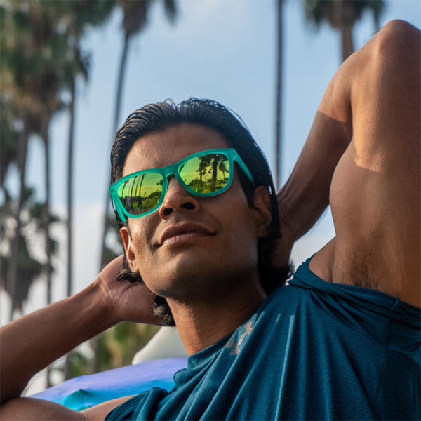 Man wearing Turquoise Goodr OG sunglasses with green lenses with palm trees in the background.