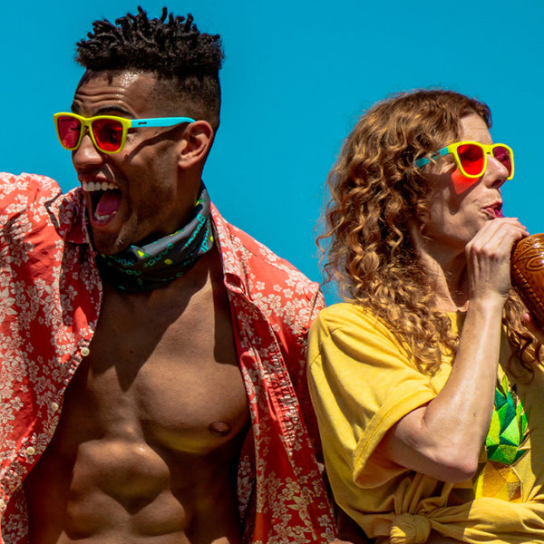 Two people wearing colorful Goodr OG Sunglasses with blue and yellow frame and pink lenses against a blue sky.