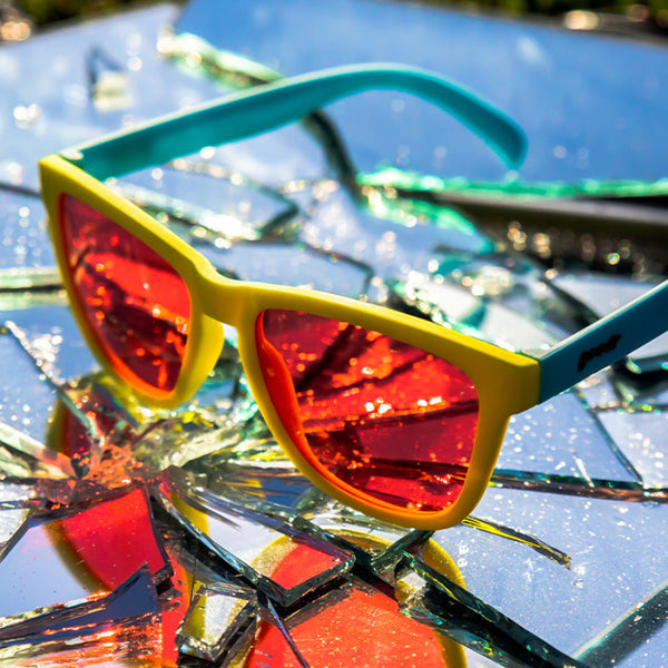 Goodr OG Sunglasses with blue and yellow frame and pink lenses on a glass surface..