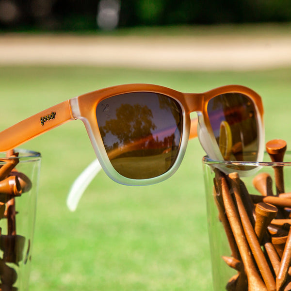 Goodr OG Three Parts Tee Sunglasses with a brown frame sitting on cups of golf tees with a blurred grassy background.