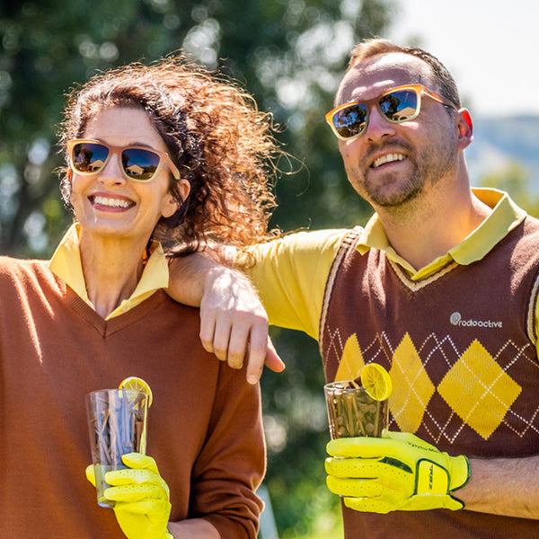 Two people wearing Goodr OG Three Parts Tee Sunglasses with a brown frame and black lenses on a blurred outdoor background.