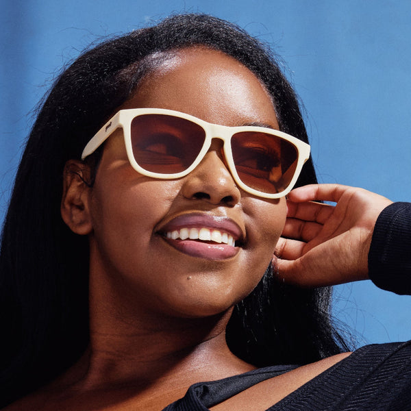 Woman wearing beige Goodr OG Toasted Marshmallow Magic sunglasses with brown gradient lenses with a clear blue sky background.