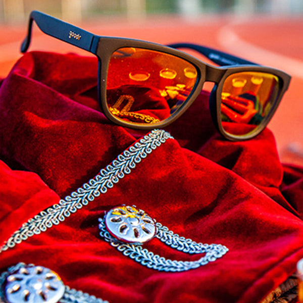 Goodr OG Whiskey Shots With Satan Sunglasses with black frame on a red fabric item with blurred background.