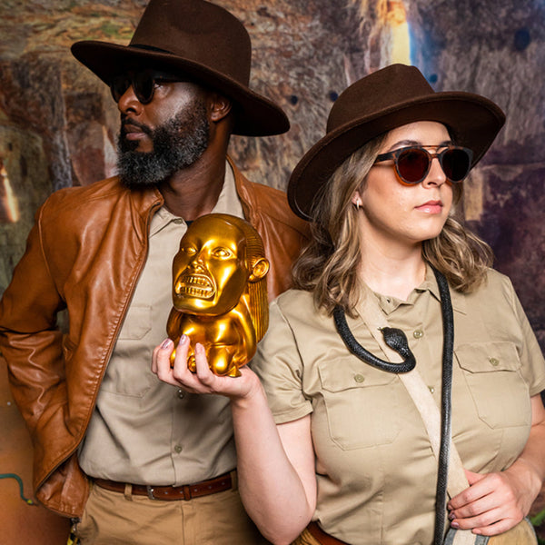 Two people in safari outfits with a gold mask and Goodr PHG sunglasses against a textured wall.