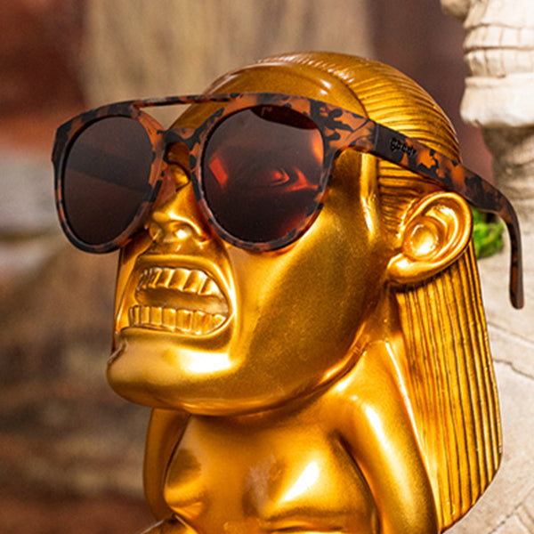 Gold statue wearing Goodr PHG Sunglasses with tortoiseshell frame and brown lenses on a blurred background.