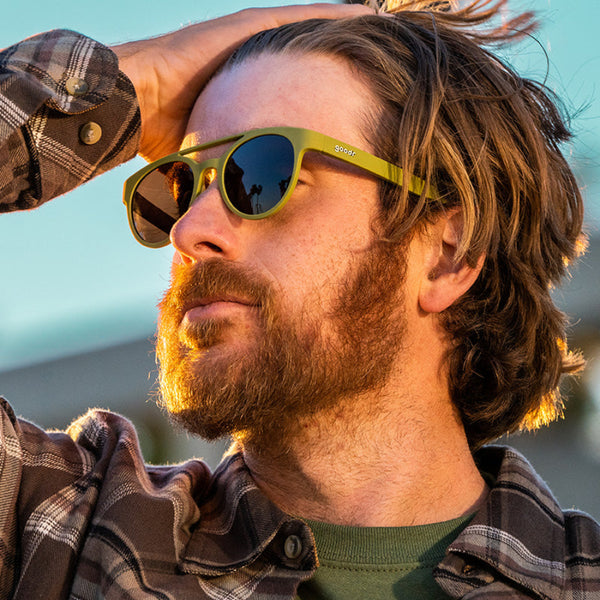 Person wearing Green Goodr Fossil Finding Focals PHG sunglasses on a blurred background.