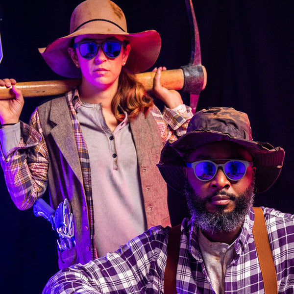 Two people dressed as prospectors  wearing Goodr PHG Sunglasses with pink lenses and dark grey frames in front of a purple curtain.