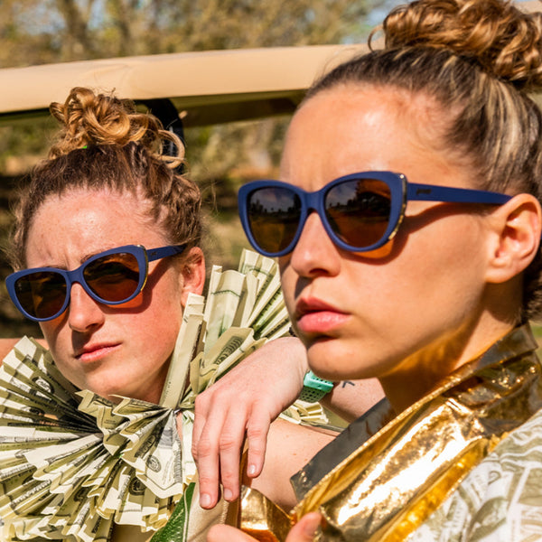 Two people wearing gold clothing and Blue Goodr Runway Mind the Wage Gap sunglasses with a gold details on a white background.