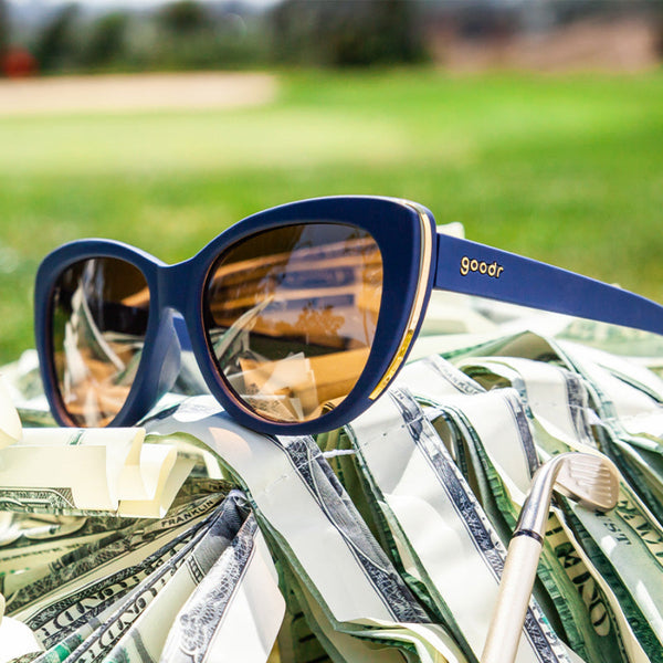 Blue Goodr Runway Mind the Wage Gap sunglasses with a gold details on  a pile of money with a grassy background.