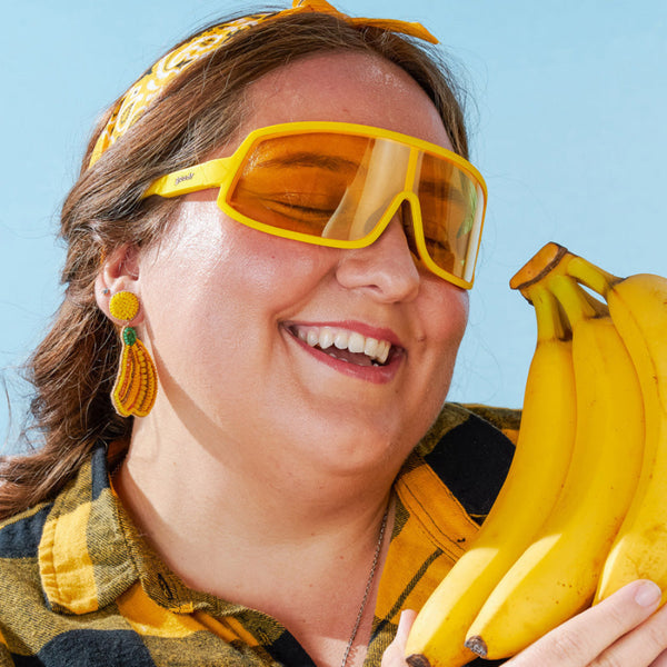 Person wearing Goodr Wrap G sunglasses and holding a bunch of yellow bananas against a blue sky background.