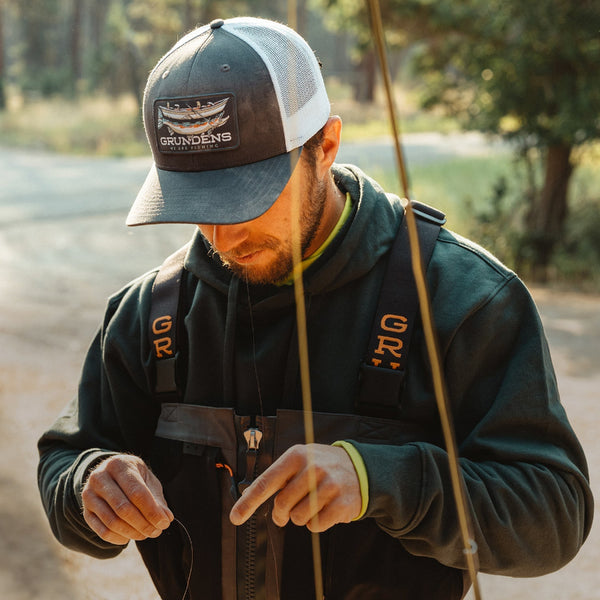 A person wearing fishing waders and a Grundéns trucker hat setting up a fly fishing rod, with a river in the background.