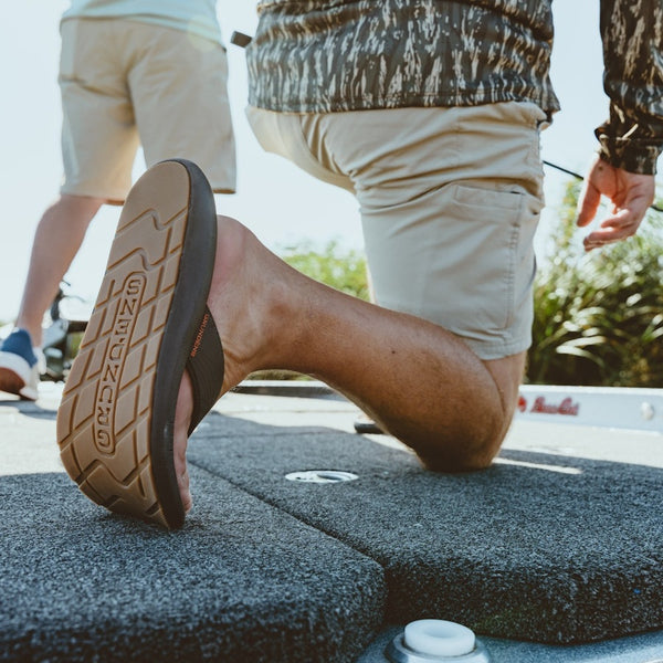 Person wearing dark brown Grundens Men's All Waters Sandals kneeling on a boat deck by a body of water with greenery in the background.