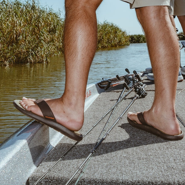 Person wearing dark brown Grundens Men's All Waters Sandals standing on a boat deck by a body of water with greenery in the background.