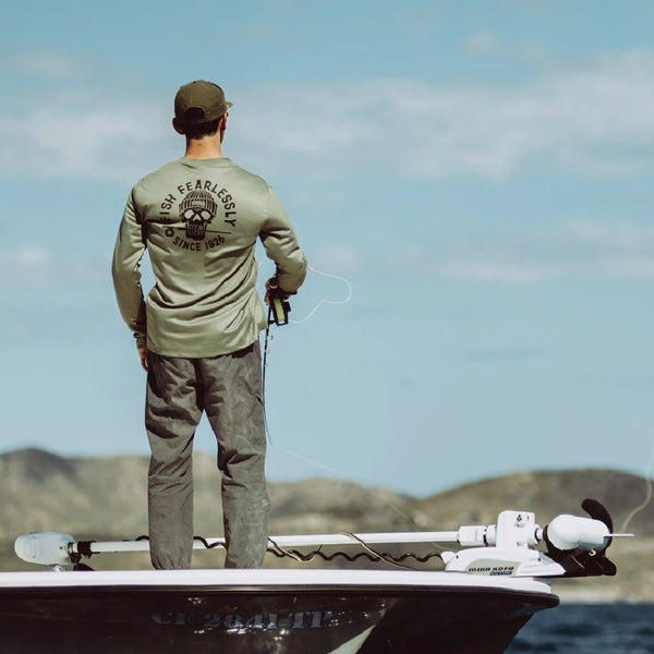 Man fly fishing on a boat wearing a forest green Grundens DeadStick Tech Tee with the shoreline in the background.