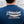 Upper back and neck of a man wearing the Grundens Men's Drift Boat Long Sleeved Tech Tee in blue abyss, with the drift boat graphic in the center, looking out over a body of water.