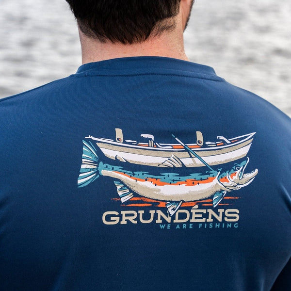 Upper back and neck of a man wearing the Grundens Men's Drift Boat Long Sleeved Tech Tee in blue abyss, with the drift boat graphic in the center, looking out over a body of water.