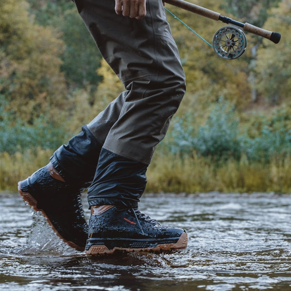 Person wearing waders and Grundens Men's Rock Armor Wading Boots walking through water with a flyfishing rod.
