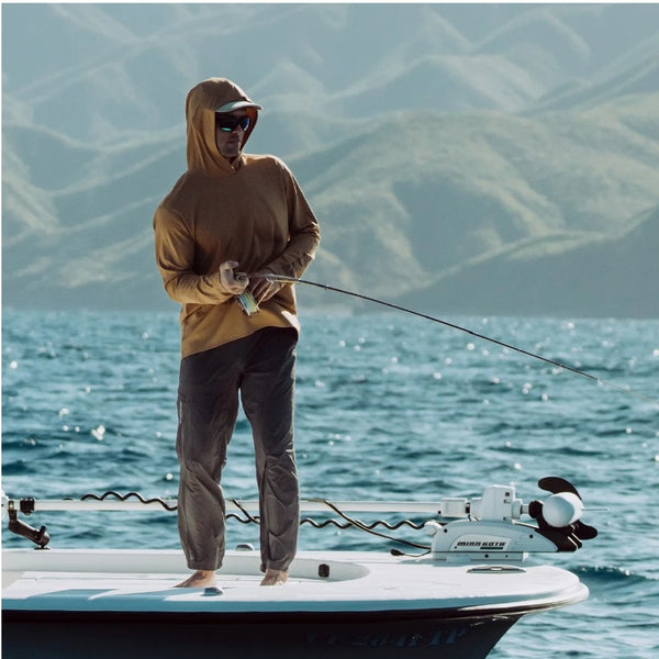 A man wearing the whiskey-colored Grundens Men's Tough Sun Hoody, fly fishing off a modern boat deck on a lake.