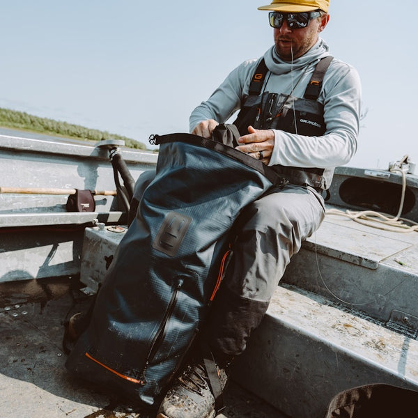 Man wearing waders sitting on a boat with fishing gear, unpacking a dark navy blue Grundéns Wayward Roll Top Backpack on a sunny day.