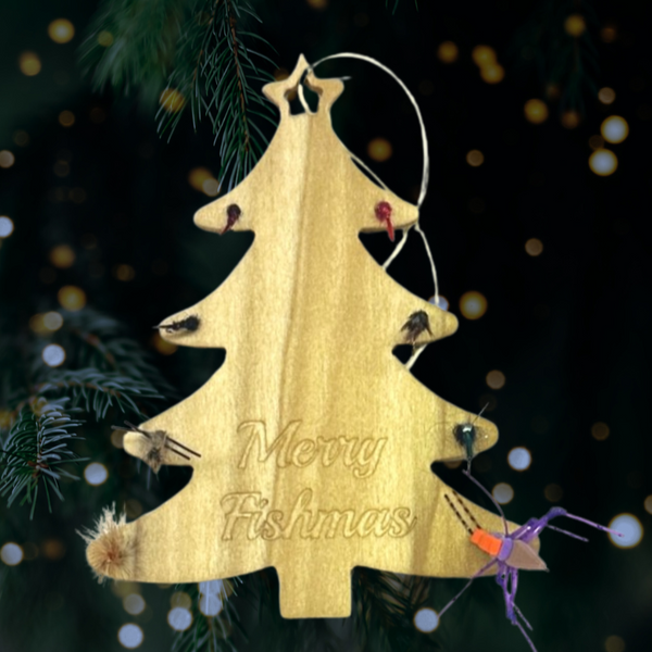 A tree shaped wooden ornament with trout flies around the edge and "Merry Fishmas" engraving over an evergreen tree with lights on the background.