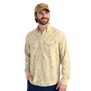 Man wearing a Huk KC Sand Camo Long-Sleeve Tide Point Shirt and brown cap on a white background.