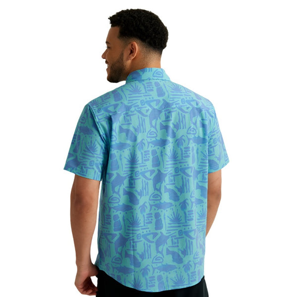 back view of a man wearing a tiki beach print marine blue color huk fishing men's kona button down short sleeve shirt on a white background