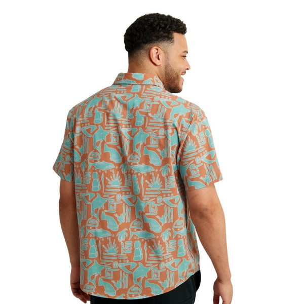 back view of a man wearing a tiki beach print sunburn color huk fishing men's kona button down short sleeve shirt on a white background