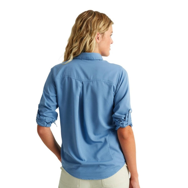 back view of a woman wearing a light blue huk fishing womens tide point button down long sleeve shirt and khaki pants on a white background