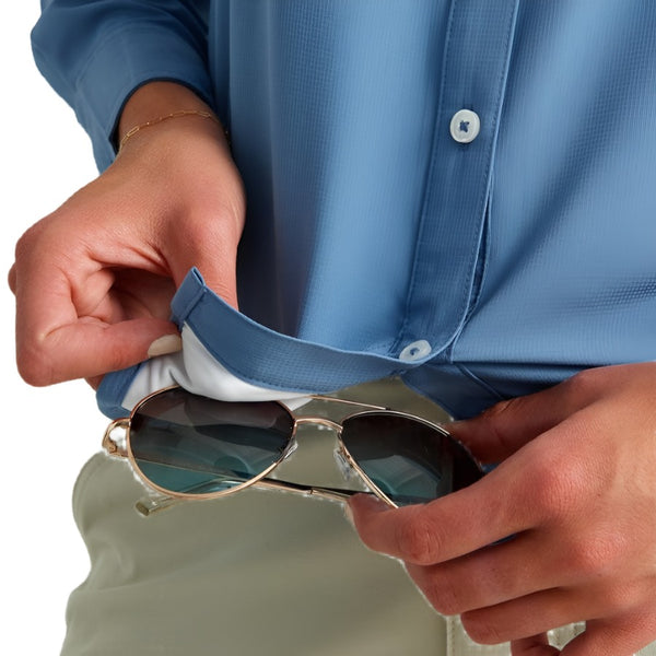 a woman cleaning aviator sunglasses with the built in lens cloth of the light blue huk fishing womens tide point button down long sleeve shirt she is wearing on a white background