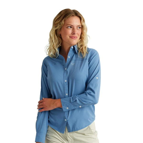 front view of a woman wearing a light blue huk fishing womens tide point button down long sleeve shirt and khaki pants on a white background