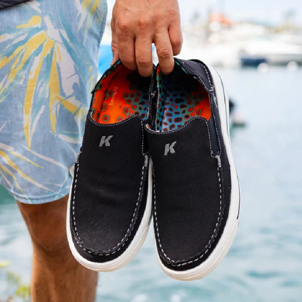 Person carrying a pair of black slip-on Korkers Men's Fish Moc shoes showing the brown trout interior, on a beachy blurred background.