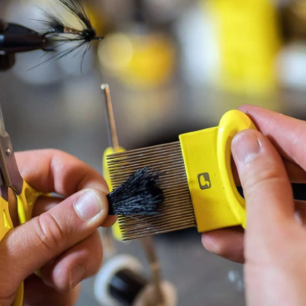 Person using a Loon Outdoors Ergo Underfur Comb fly tying tool to remove the underfur from a clump of hair with a tying vise and tools blurred in the background.