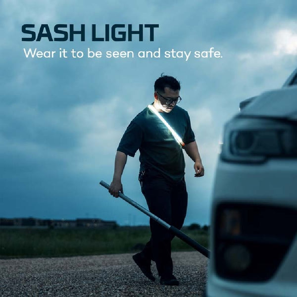 Person wearing the Nebo Bando 1k work light as a sash light on a road with a vehicle in the foreground and text 'Wear it to be seen and stay safe.'.
