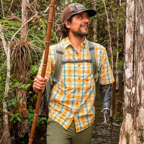 A man wearing a backpack, the Orvis Men's Open Air Caster Short Sleeved Plaid Shirt in tangerine/peacock, and a prosthetic arm hikes through the Florida Everglades with a bamboo walking stick. 