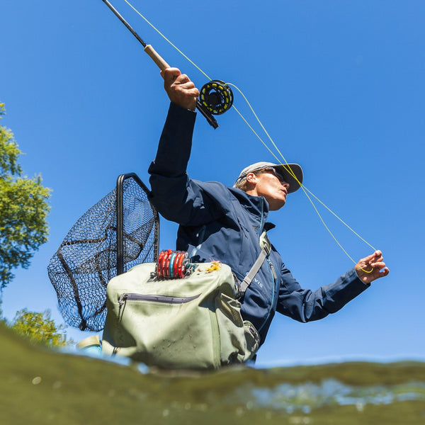 Person standing in the water casting a fly fishing rod against a cleat blue sky and wearing the Orvis Pro Waterproof Hip Pack fully loaded with tippet, flies and a landing net in the integrated net scabbard.