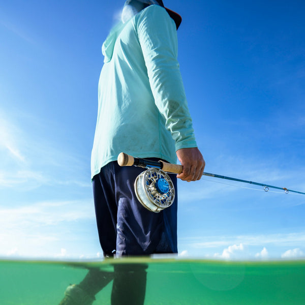Person holding a fishing rod with the Orvis Ratio Saltwater Fly Reel in silver, with a blue sky and water background.