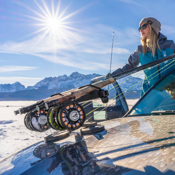 Person with Orvis Recon fly fishing rods on a car hood rack in a snowy landscape with mountains.