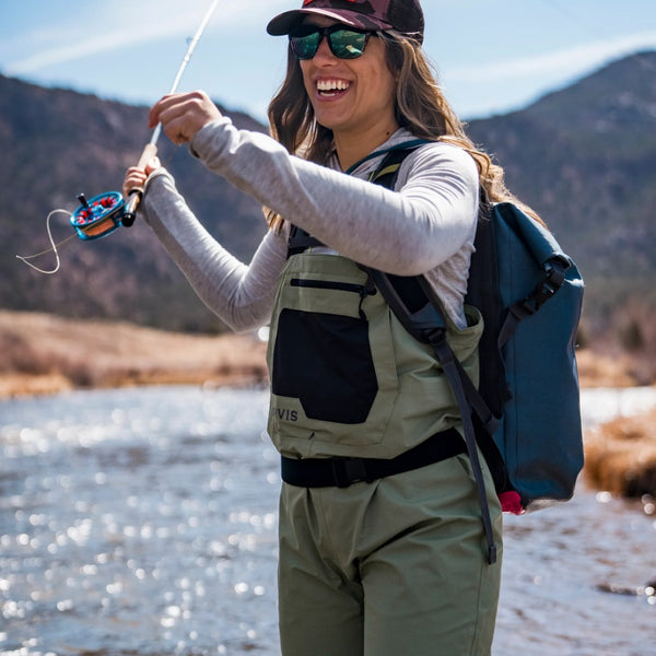 A woman fly fishing in a mountain stream wearing a set of Orvis Women's Clearwater Stockingfoot Waders in sagebrush and a blue backpack, against a blurred natural background.