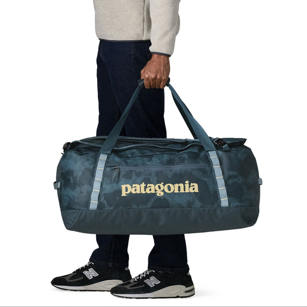 Person holding the straps of the sastrugi summit blue 70-Liter Patagonia Black Hole Duffel Bag showing side logo and pocket, on a white background.