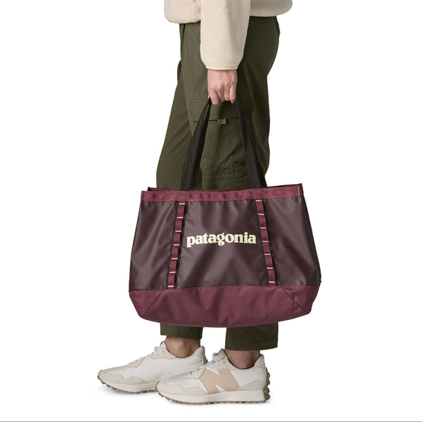A person in green pants and sneakers carrying the 25-liter Patagonia Black Hole Gear Tote in berry fig, on a white background.