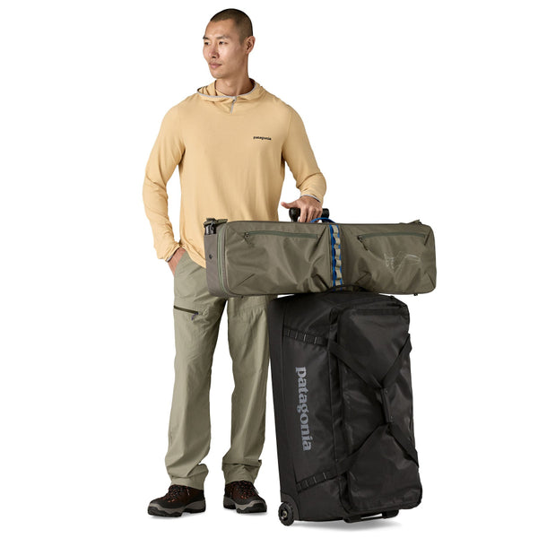 Man holding a basin green Patagonia Black Hole Fly Fishing Rod Case on top of a Black Hole rolled duffel, on a white background.