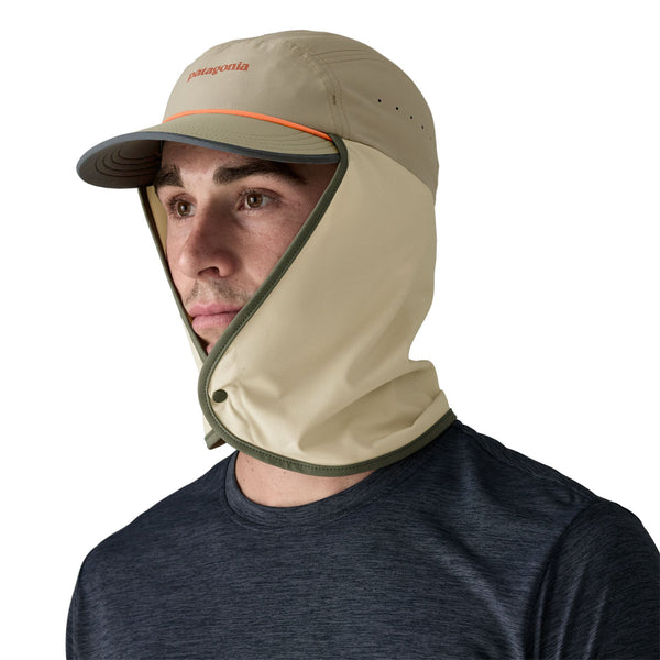 Angled front view of a person wearing a dark blue shirt and the river rock green Patagonia Caped Merganzer Hat with the full neck coverage panel snapped in front, against a white background.