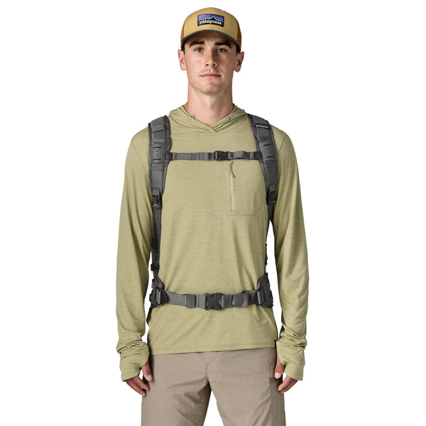 A person shown from the front wearing a light green sun hoody and the forge grey 25-Liter Patagonia Guidewater Waterproof Backpack on a white background.