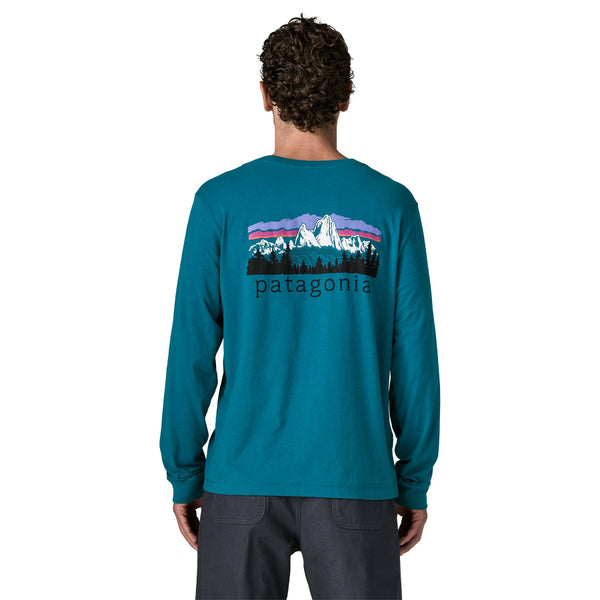 A person wearing black pants and a glacial blue color Patagonia Men's Fitz Roy Foothills Long-Sleeved T-Shirt showing the large logo graphic on the back, against a white background.