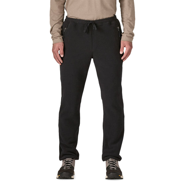 A person wearing a tan long sleeved shirt and lack solid color Patagonia Men's Synchilla Fleece Pants with an elastic waistband, drawstring, and zippered side pockets, against a white background.