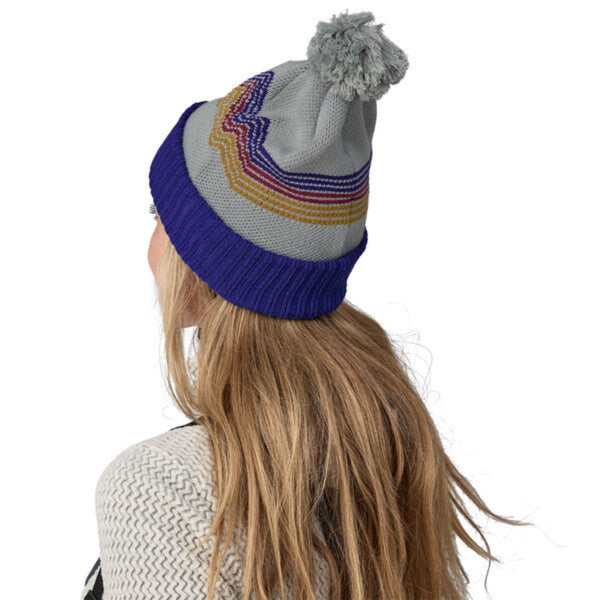 Person wearing a knitted Patagonia Branded beanie in grey with colorful pattern, pom pom topper, and blue band on a white background.