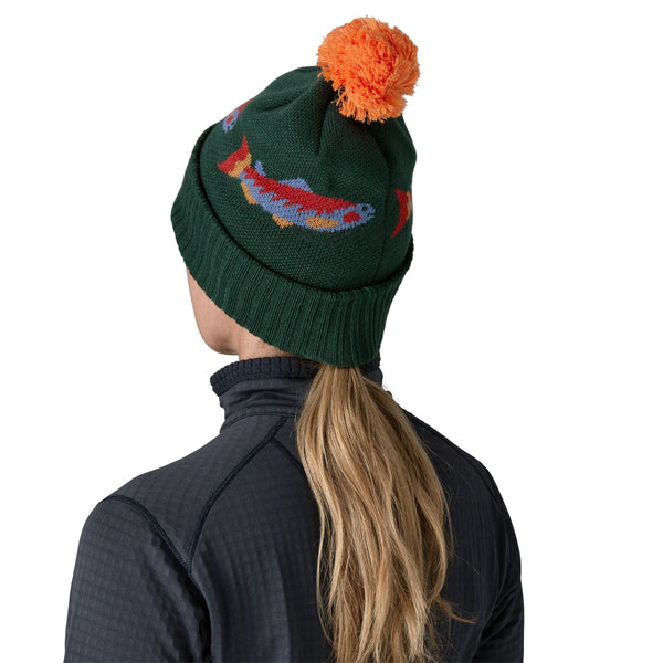 Person wearing a green Powder Town Knit beanie with a colorful pom-pom and salmon designs, and a dark jacket with a Patagonia logo over a white background.